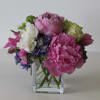 Low arrangement of pink peonies, white and green roses, and purple blooms in a clear glass cube vase