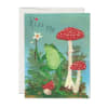Folded greeting card with a frog holding a white flower among red mushrooms and the words “Kiss Me.”