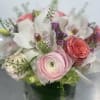 Short arrangement of pink ranunculus, roses, white orchids, and purple blooms in a clear glass cube vase