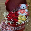 Red hat box filled with red roses, gold-wrapped chocolates, and a white teddy bear holding a heart.