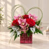 Pink orchids and red roses in a red glass vase with arched grass loops