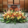 Low, wide arrangement of yellow lilies and peach flowers with large green foliage in front of a stage podium