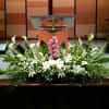 Large white and green altar arrangement with lilies, calla lilies, and a pink flower cluster beneath a green cross.