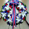 Large floral wreath with white lilies, red carnations, blue flowers, and a tricolor ribbon bow