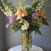 Tall mixed bouquet with peach roses, white blooms, lilac stems in a clear glass cylinder vase.