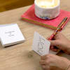 Hands writing in a floral greeting card beside a lit candle and card box