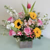 Mixed bouquet with pink roses, sunflowers, and white filler flowers in a square vase
