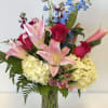 Bouquet of pink lilies, red roses, blue delphinium, and pale hydrangeas in a glass vase