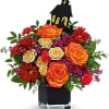 Orange and red roses with red daisies in a black vase topped by a haunted house decoration