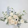 Low arrangement of ivory roses, white blooms, and pale blue flowers in a white ceramic vase