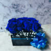 Low black box arrangement of vivid blue roses with white filler flowers and a large blue ribbon bow.