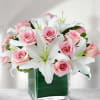 Pink roses and white lilies in a clear glass cube vase lined with a green leaf.