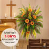 Tall sympathy easel spray with orange gerbera daisies, yellow blooms, and ferns beside a casket.