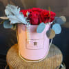 Red roses arranged in a pink hatbox with pearl strands and blue-gray foliage.