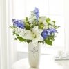 White lilies and blue-white flowers arranged in a tall vase with a cross emblem.