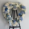 Heart-shaped floral wreath with white roses and blue hydrangeas on a black stand