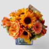 Compact bouquet of yellow sunflowers, orange roses, and peach gerberas in a square glass vase