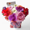 Compact arrangement of pink roses, purple orchids, and mixed blooms in a square container with butterfly decoration.