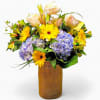 Mixed bouquet of yellow daisies, blue hydrangeas, and pale roses in a tall burlap-wrapped vase