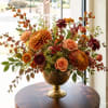 Orange and burgundy floral arrangement in a gold pedestal vase