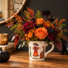 Orange and burgundy floral arrangement in a decorated mug