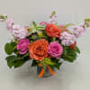 Short arrangement of orange and pink roses with pale pink stock and a succulent in a metal vase.