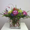 Pink and purple roses arranged in a clear square vase with curly wire accents