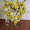 Heart-shaped standing wreath of yellow roses, white daisies and lilies with a white bow on an easel.