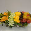 Long floral centerpiece with orange lilies, yellow roses, and mixed blooms in a rectangular vase