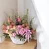 Pink and cream floral arrangement in a white bowl vase with a lavender ribbon