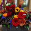 Colorful floral arrangement with roses, gerbera daisies, sunflowers, and a small pumpkin in a metal container
