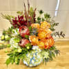Mixed bouquet with orange roses, blue hydrangea, and burgundy protea in a low vase