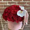 Round pink box of red roses topped with white orchids and pink ribbons against a brick wall.