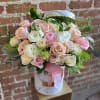 Round hatbox of blush and cream roses with white blooms and greenery against a brick wall