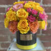 Compact arrangement of yellow, pink, and peach roses in a black hatbox-style container with a yellow band