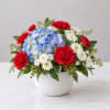 Red carnations, blue hydrangea, and white daisies in a white bowl vase