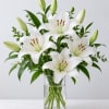 White lilies arranged in a clear glass vase with green buds and foliage