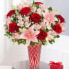 Red roses and pink lilies in a red glass vase beside a small gift box
