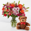 Mixed bouquet in a glass vase beside a teddy bear with a red bow