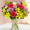 Bouquet of pink roses, yellow lilies, and lavender daisies in a glass vase