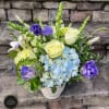 Arrangement of pale yellow roses, blue hydrangeas, and purple blooms in a white vase on stone steps