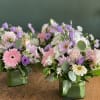 Two pastel floral arrangements in square glass vases on a wooden table