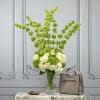 Tall green and white floral arrangement in a clear vase beside a beige handbag
