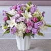 Tall arrangement of white lilies, pink roses, and lavender flowers in a white vase