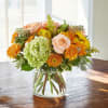 Round arrangement of peach roses, green hydrangea, and orange mums in a clear glass vase