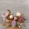 Pastel pink and peach floral arrangement with lavender mums and autumn leaves in a white vase on a woven basket.