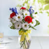 Bouquet of white daisies, red carnations, and blue flowers in a glass vase with a yellow ribbon
