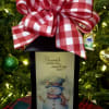 Gift box with a red plaid bow and a snowman card in front of a Christmas tree