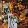 Orange and yellow flower arrangement in a white lantern-style holder