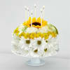 Round floral cake of white daisies, yellow blooms, and a sunflower on a stand with six lit candles on top.